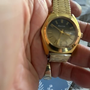 May include: A gold-toned Bulova wristwatch with a rectangular face and a mesh band. The watch face is gold with black accents and displays the day and date. The watch is held in a hand.