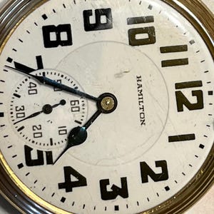 May include: A gold pocket watch with a white face and black Roman numeral hour markers. The watch has a small second hand dial at the 3 o'clock position. The watch face also has the word "HAMILTON" printed on it.