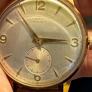 Puede incluir: Un reloj de pulsera dorado con una esfera blanca y agujas negras. La esfera del reloj tiene marcadores de hora romanos negros y un pequeño dial de segundos en la posición de las 6 en punto. La esfera del reloj también tiene el texto "Aureola 19 Rubis".