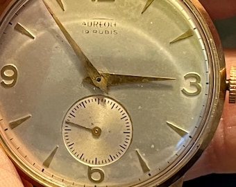 Puede incluir: Un reloj de pulsera dorado con una esfera blanca y agujas negras. La esfera del reloj tiene marcadores de hora romanos negros y un pequeño dial de segundos en la posición de las 6 en punto. La esfera del reloj también tiene el texto "Aureola 19 Rubis".