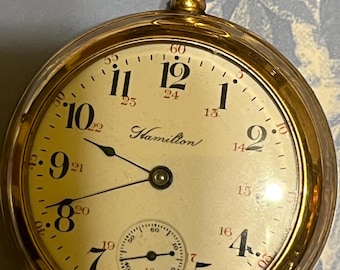 Puede incluir: Un reloj de bolsillo Hamilton dorado con esfera crema. Números negros marcan las horas, con un pequeño dial de segundos. El reloj tiene una corona dorada y un lazo para una cadena. La palabra "Hamilton" está impresa en la esfera.