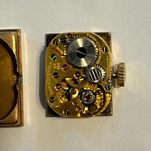 K&ouml;nnte beinhalten: Nahaufnahme eines Vintage-Uhrwerks mit komplizierten Zahnr&auml;dern, Juwelen und einer Aufzugswelle. Der goldfarbene Mechanismus ist rechteckig, mit sichtbarem Text und einer kleinen, runden Krone. Das Uhrwerk liegt auf einer wei&szlig;en Oberfl&auml;che.