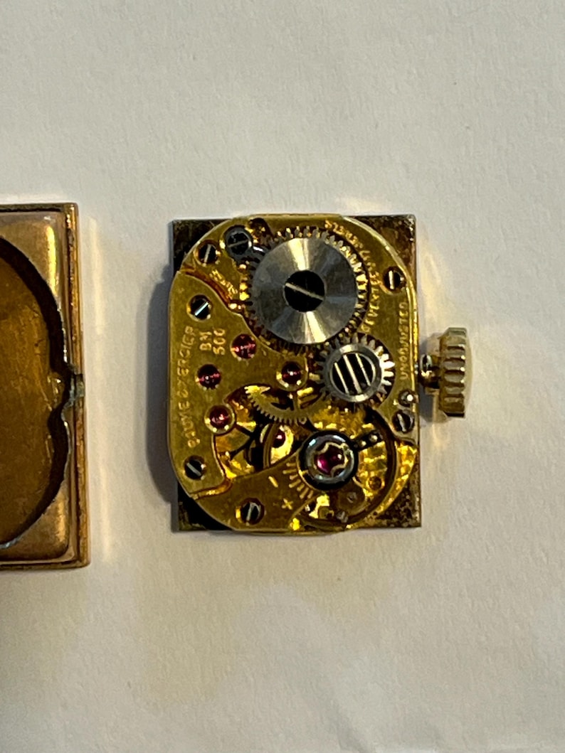 K&ouml;nnte beinhalten: Nahaufnahme eines Vintage-Uhrwerks mit komplizierten Zahnr&auml;dern, Juwelen und einer Aufzugswelle. Der goldfarbene Mechanismus ist rechteckig, mit sichtbarem Text und einer kleinen, runden Krone. Das Uhrwerk liegt auf einer wei&szlig;en Oberfl&auml;che.