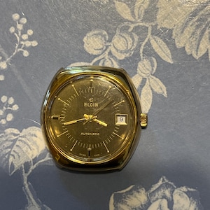 May include: A vintage Elgin automatic wristwatch with a gold-toned case and a dark face. The watch features gold hands and hour markers, a date window at the 3 o'clock position, and the word "ELGIN" at the top. The watch is set against a floral background.
