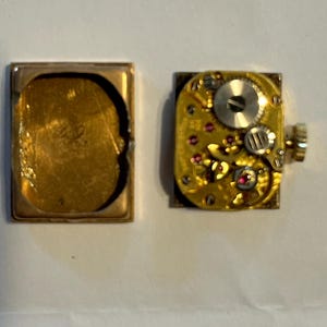 K&ouml;nnte beinhalten: Eine zerlegte Vintage-Uhr, die ihr Innenleben zeigt. Das goldfarbene Geh&auml;use ist ge&ouml;ffnet und gibt den Blick auf das komplizierte Uhrwerk mit kleinen Zahnr&auml;dern, Juwelen und einer Aufzugswelle frei. Das Uhrwerk der Uhr ist quadratisch.