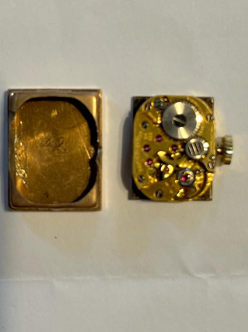 K&ouml;nnte beinhalten: Eine zerlegte Vintage-Uhr, die ihr Innenleben zeigt. Das goldfarbene Geh&auml;use ist ge&ouml;ffnet und gibt den Blick auf das komplizierte Uhrwerk mit kleinen Zahnr&auml;dern, Juwelen und einer Aufzugswelle frei. Das Uhrwerk der Uhr ist quadratisch.