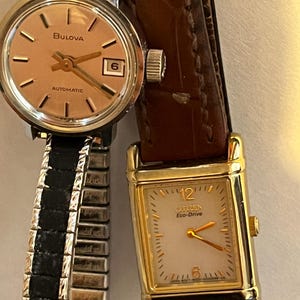 May include: Two wristwatches. The first is a Bulova automatic watch with a round, gold-tone face and a black and silver band. The second is a gold-tone Citizen Eco-Drive watch with a rectangular face and a brown leather band.