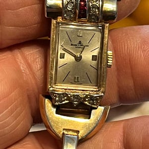 K&ouml;nnte beinhalten: Vintage goldfarbene Baume & Mercier Geneva Uhr. Das rechteckige Zifferblatt hat ein silbernes Zifferblatt mit schwarzen Markierungen und Zeigern. Die Uhr hat ein goldfarbenes Armband mit dekorativer Oberseite und ist mit kleinen klaren und roten Edelsteinen verziert.