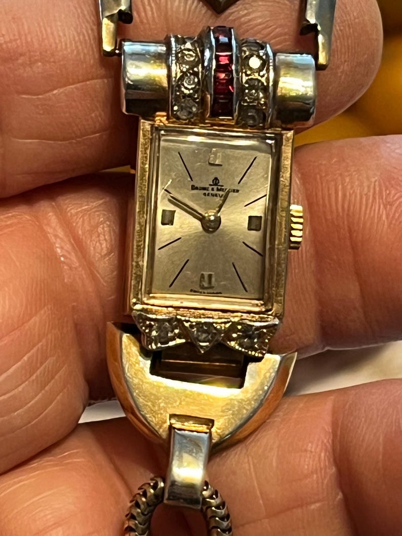 K&ouml;nnte beinhalten: Vintage goldfarbene Baume & Mercier Geneva Uhr. Das rechteckige Zifferblatt hat ein silbernes Zifferblatt mit schwarzen Markierungen und Zeigern. Die Uhr hat ein goldfarbenes Armband mit dekorativer Oberseite und ist mit kleinen klaren und roten Edelsteinen verziert.