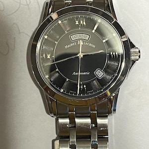 May include: A silver-toned Maurice Lacroix automatic wristwatch. The watch face is dark gray with silver hands and hour markers. The day of the week, "Saturday", is displayed above the brand name. The watch has a date display at the 3 o'clock position and a stainless steel band.