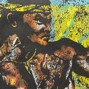 May include: Close-up of a black t-shirt featuring a distressed graphic of a person's face with a yellow and blue background. The graphic has a vintage, worn look with brown and white accents.