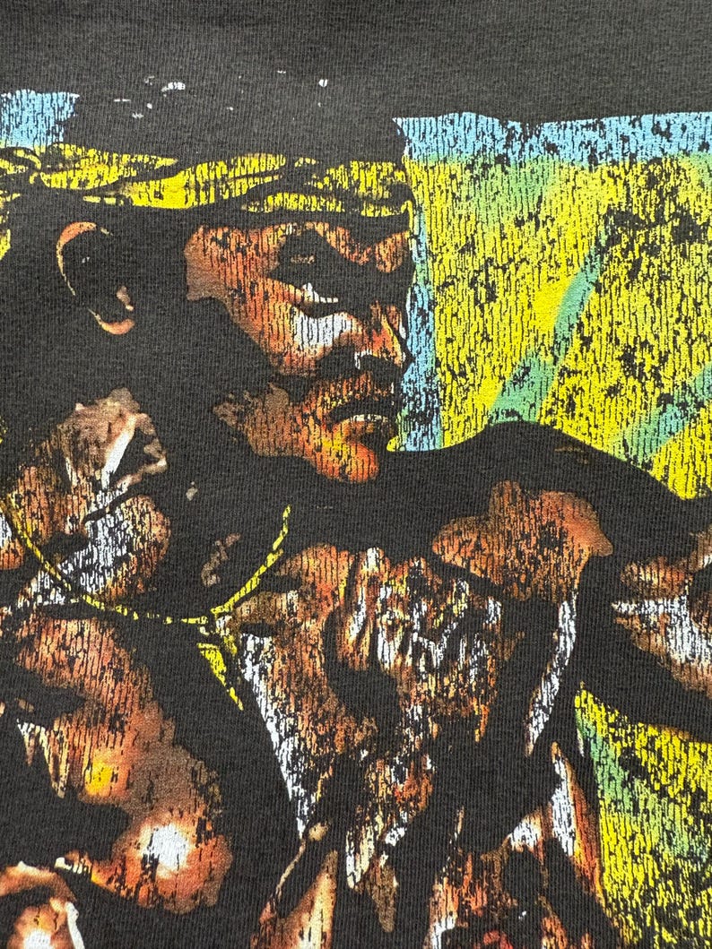May include: Close-up of a black t-shirt featuring a distressed graphic of a person's face with a yellow and blue background. The graphic has a vintage, worn look with brown and white accents.