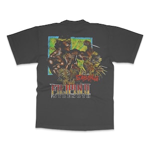 May include: Dark gray t-shirt featuring a graphic of two muscular figures in combat, with a knife. The design includes the text "SABONG" and "FILIPINO STRENGTH" in a vintage style, with a blue, green, and yellow background.