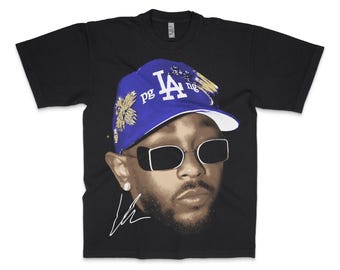 Op de afbeelding: Zwart T-shirt met een afbeelding van een persoon die een blauwe en gouden baseballpet van de Los Angeles Dodgers draagt en een zonnebril. De afbeelding bevat de tekst "pg" en een handtekening.