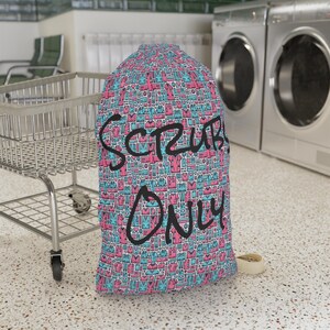 Könnte beinhalten: Eine blaue und rosafarbene Wäschetasche mit weißem Hintergrund und einem Muster aus verschiedenen Kleidungsstücken. Die Tasche trägt die Aufschrift "Scrubs Only" in Schwarz auf der Vorderseite.