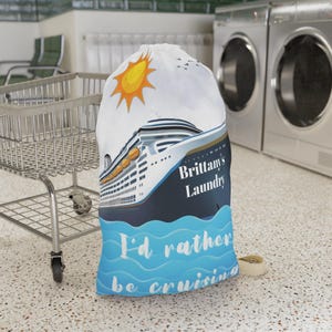 May include: White laundry bag with a blue and white cruise ship, a yellow sun, and the text "Brittany's Laundry I'd rather be cruising".