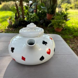 May include: A white ceramic teapot featuring a repeating pattern of black and red playing card suit symbols, including hearts, diamonds, clubs, and spades. The teapot has a rounded body, a short spout, and a matching white lid with a knob handle.
