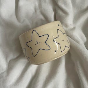 May include: A cream-colored ceramic mug featuring hand-drawn dark blue star shapes with simple facial expressions. The surface is decorated with small dots and tiny star accents scattered between the larger star illustrations on the exterior.