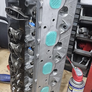 May include: A silver engine cylinder head with a series of blue valve seals. The cylinder head is resting on a yellow cloth. A can of WD-40 is visible in the background.