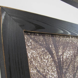 May include: Close-up of a black wood picture frame with a visible wood grain. The frame is surrounding a print of a tree with brown leaves.