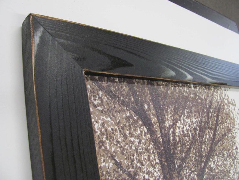 May include: Close-up of a black wood picture frame with a visible wood grain. The frame is surrounding a print of a tree with brown leaves.