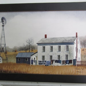 May include: A framed painting of a white farmhouse with a windmill in the foreground. The house has a whitewashed exterior with blue window frames and a red brick chimney. The windmill is made of metal and has a wooden base. The painting is set against a backdrop of a field with hay bales and trees. The title of the painting is "Wash Day" and is signed by the artist.