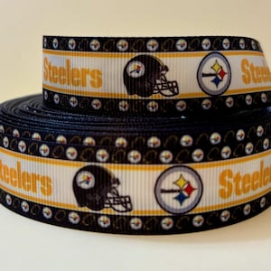 May include: A roll of black and white ribbon featuring the word "Steelers" in gold, along with football helmet and logo designs. The ribbon is suitable for crafting and sports-themed projects.