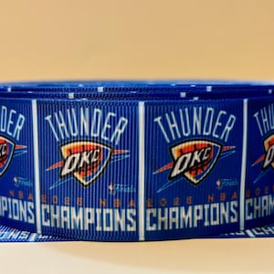 May include: A roll of blue ribbon featuring the text "THUNDER" and "OKC" with a basketball logo. The ribbon also displays "2026 NBA CHAMPIONS" in white text. The ribbon is rolled up.