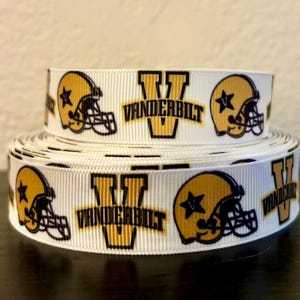 May include: White ribbon featuring a repeating pattern of gold football helmets with a star and the word "VANDERBILT" in gold and black. The ribbon is rolled up in a stack.