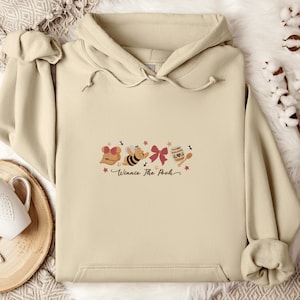May include: A beige hoodie featuring a graphic of Winnie the Pooh characters, including a gift, a bee, a bow, and a honey pot. The text "Winnie The Pooh" is written in a script font below the graphic. The hoodie has a hood and a front pocket.