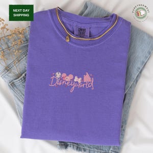 a purple t-shirt with the text "Disneyworld" printed on it, along with various Disney-themed graphics and characters. The t-shirt is folded and placed on a white surface, with a pair of jeans visible in the background.