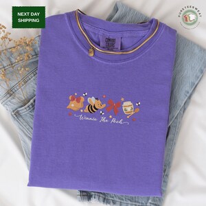May include: A purple Comfort Colors t-shirt with an embroidered design of Winnie the Pooh characters, including Pooh Bear, a bee, and a honey pot. The shirt has a gold necklace and is displayed on denim jeans.