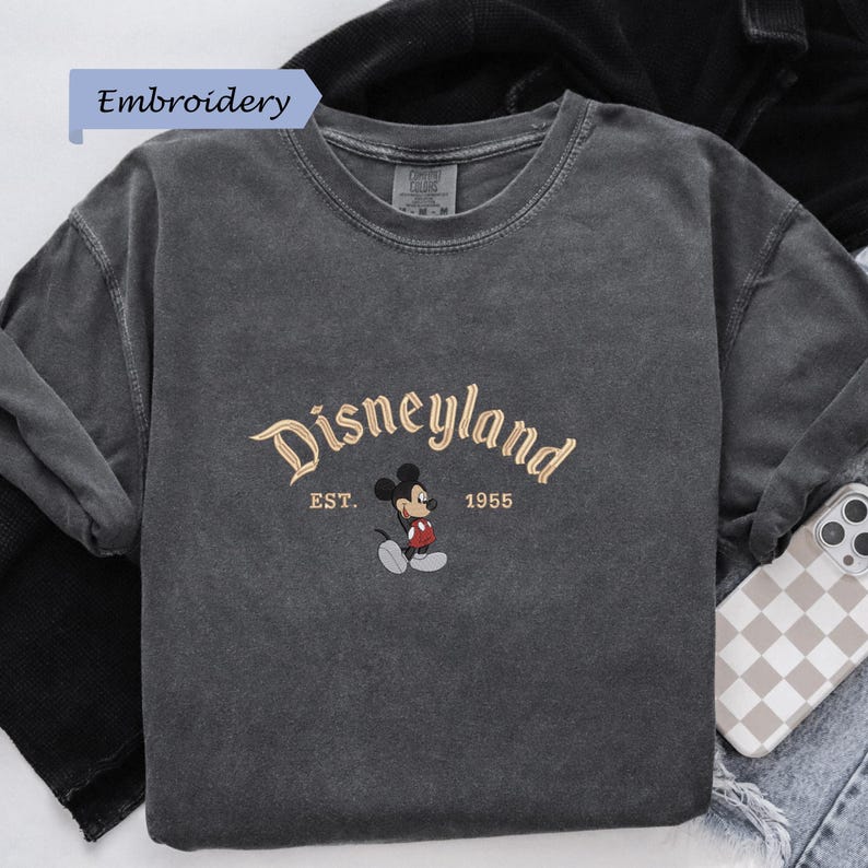 A gray t-shirt with the text "Disneyland EST. 1955" and a Mickey Mouse graphic printed on it, along with a small embroidered Mickey Mouse.