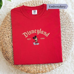 A red t-shirt with the text "Disneyland EST. 1955" and a Mickey Mouse embroidered on it, placed on a woven basket.