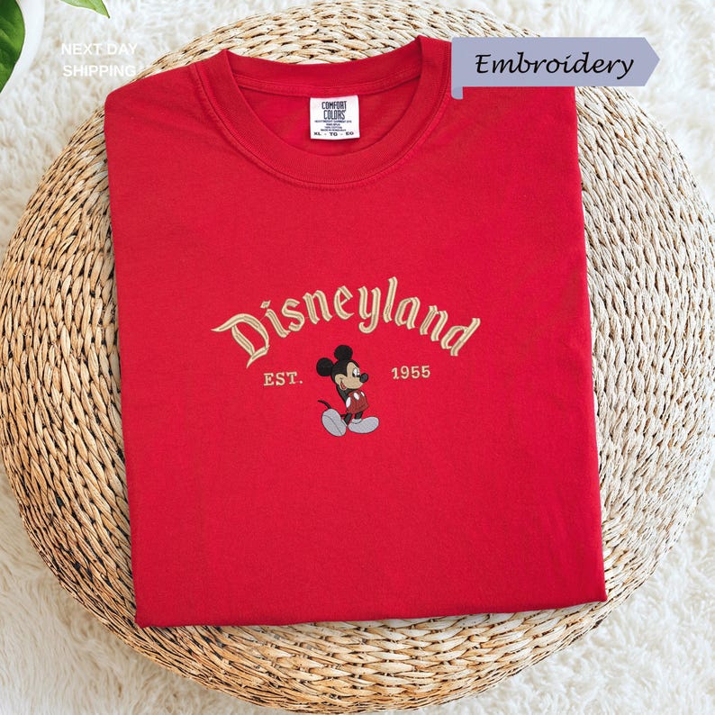 A red t-shirt with the text "Disneyland EST. 1955" and a Mickey Mouse embroidered on it, placed on a woven basket.