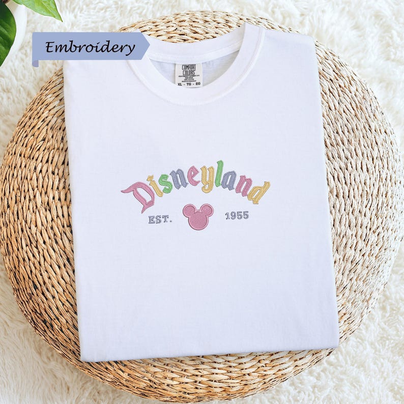 A white t-shirt with the text "Disneyland EST. 1955" embroidered on it, along with a Mickey Mouse logo.