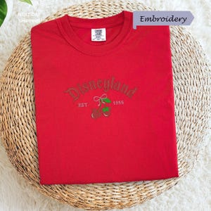 A red t-shirt with the text "Disneyland EST. 1955" printed on it, along with a small embroidered logo featuring a cherry blossom.