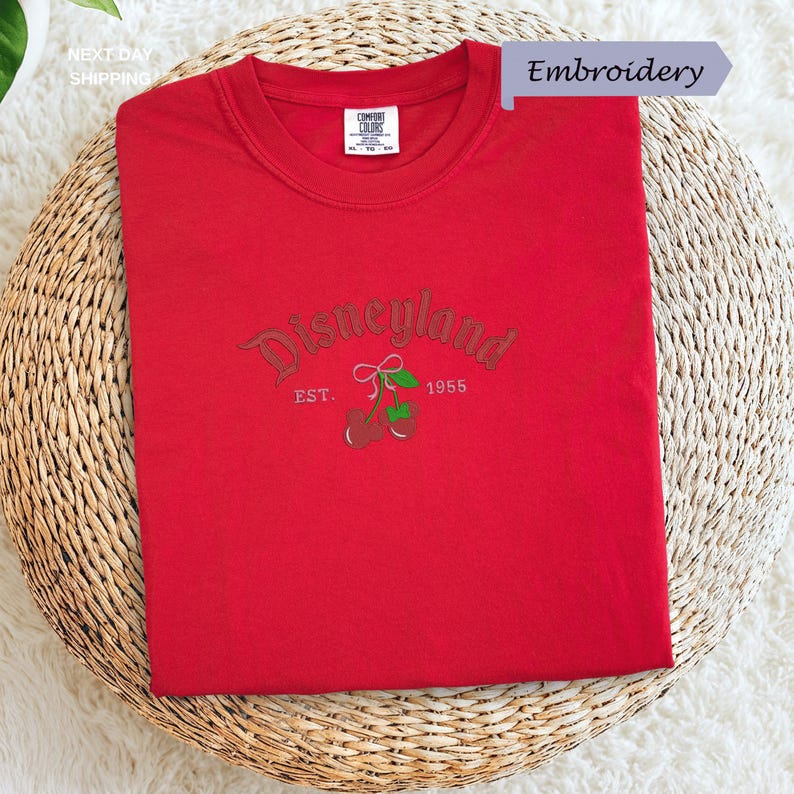 A red t-shirt with the text "Disneyland EST. 1955" printed on it, along with a small embroidered logo featuring a cherry blossom.