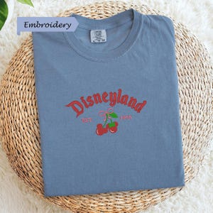 A blue t-shirt with the text "Disneyland EST. 1955" and a graphic of cherries is displayed on a woven basket.