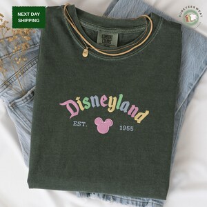 A green t-shirt with the text "Disneyland EST. 1955" printed on it, along with a Mickey Mouse logo, is displayed on a bed.