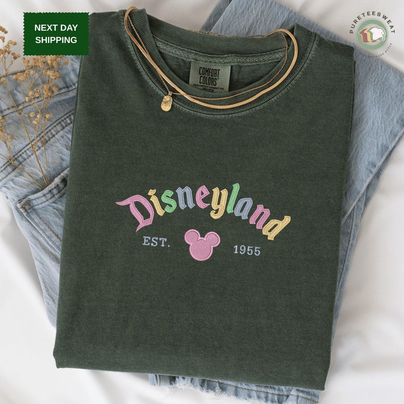 A green t-shirt with the text "Disneyland EST. 1955" printed on it, along with a Mickey Mouse logo, is displayed on a bed.