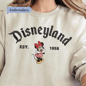 A white sweatshirt with the text "Disneyland EST. 1955" and an embroidered image of Minnie Mouse, a character from the Disney theme park.