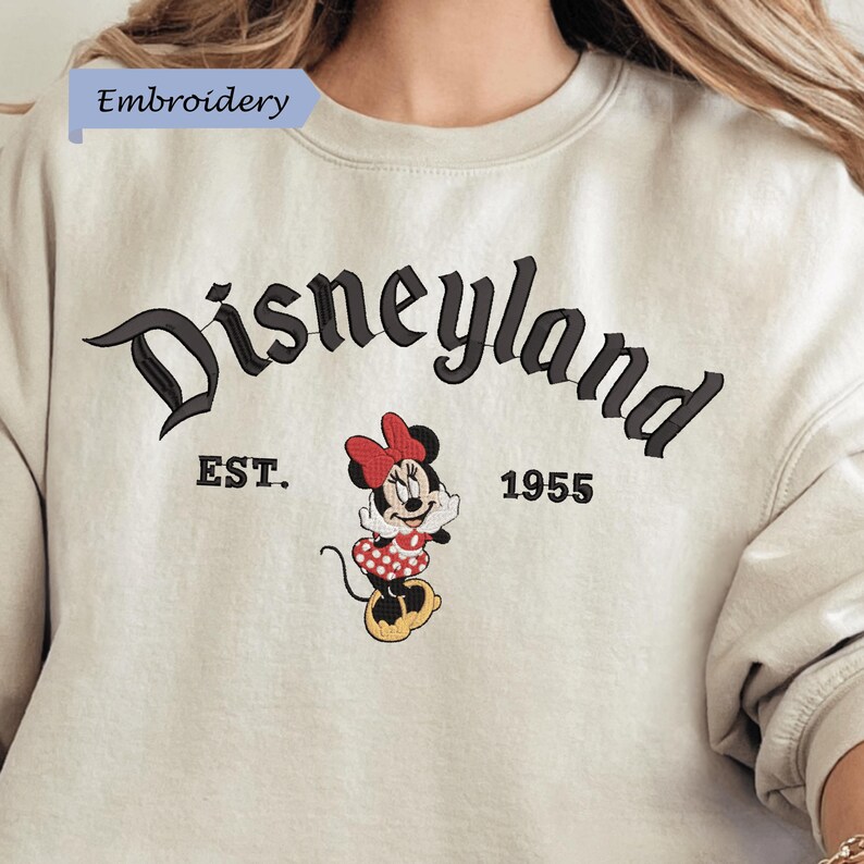 A white sweatshirt with the text "Disneyland EST. 1955" and an embroidered image of Minnie Mouse, a character from the Disney theme park.