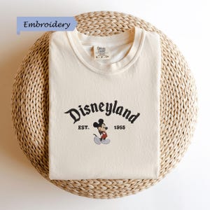 A white t-shirt with a Disney Mickey Mouse logo and text, laid out on a woven basket.