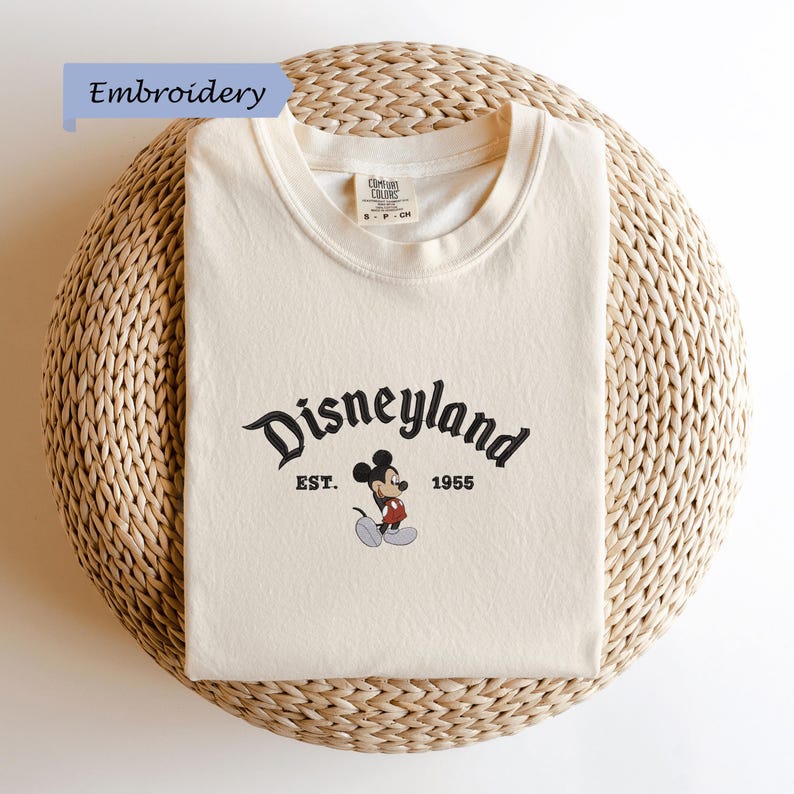 A white t-shirt with a Disney Mickey Mouse logo and text, laid out on a woven basket.
