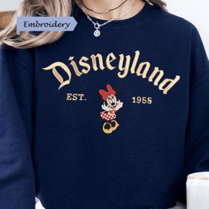 A close-up of a person wearing a navy blue sweater with the text "Disneyland EST. 1955" and an embroidered Minnie Mouse character.