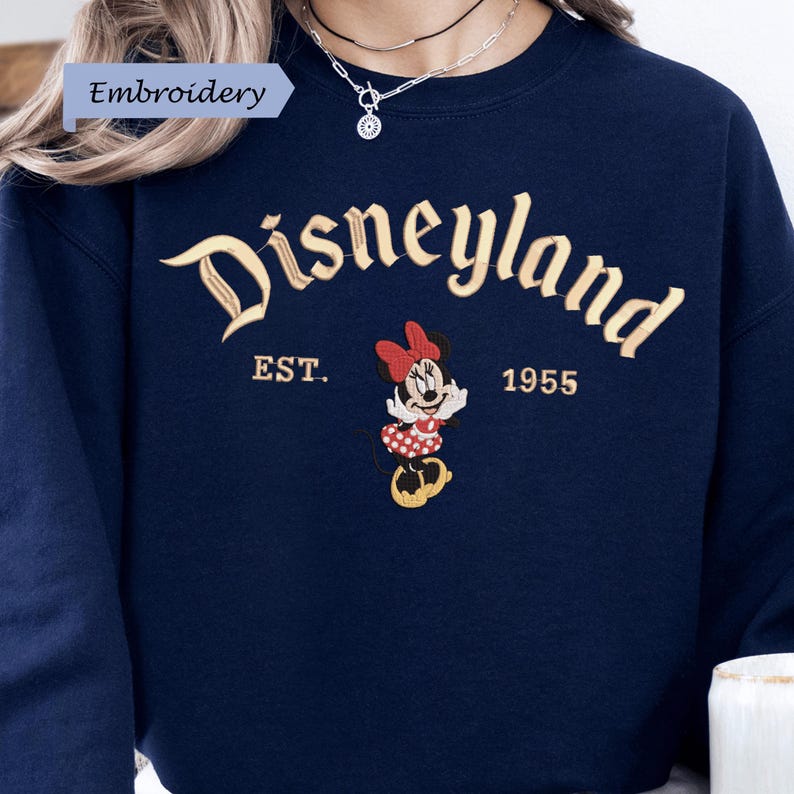 A close-up of a person wearing a navy blue sweater with the text "Disneyland EST. 1955" and an embroidered Minnie Mouse character.