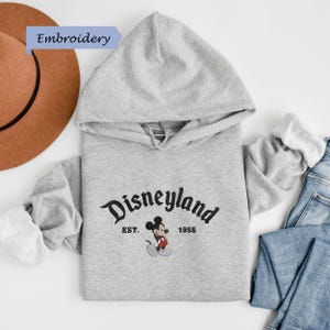 A gray hoodie with the text "Disneyland EST. 1956" printed on it, along with a Mickey Mouse logo, is laid out on a white background.