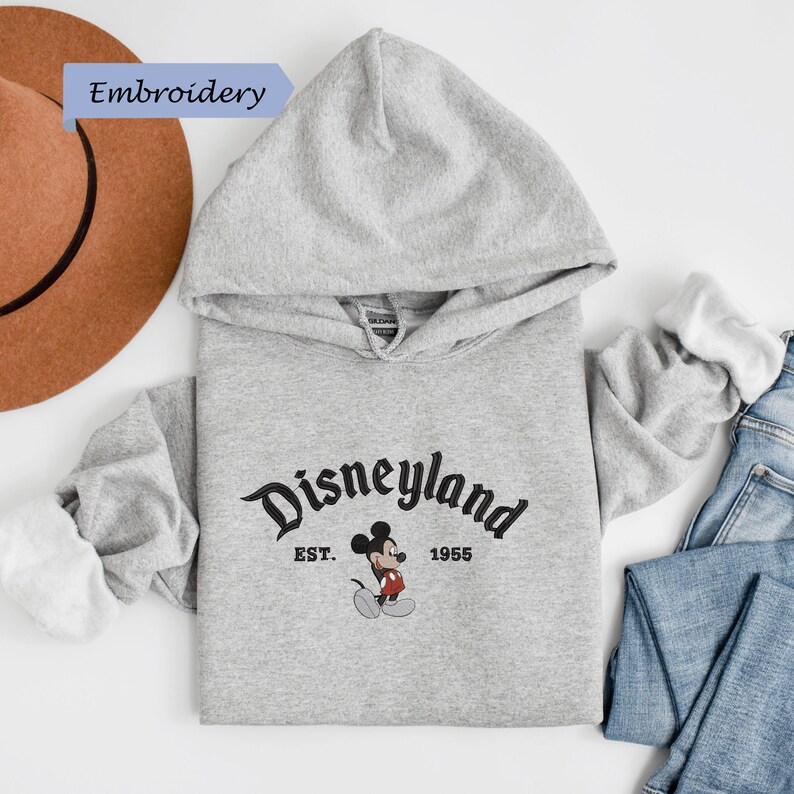 A gray hoodie with the text "Disneyland EST. 1956" printed on it, along with a Mickey Mouse logo, is laid out on a white background.
