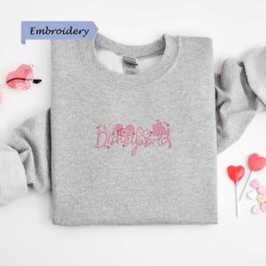 a gray sweatshirt with a pink embroidered design, along with various heart-shaped candies and a pair of sunglasses.